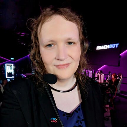 Triana wearing a broadcast headset on a stage with colourful purple and pink lights behind her. A logo for the mental health charity ReachOut is on a giant screen.